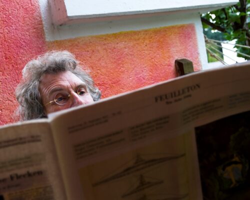 Grauhaariger Mann mit lockigem Haar blickt über die Zeitung "DerStandard" hervor