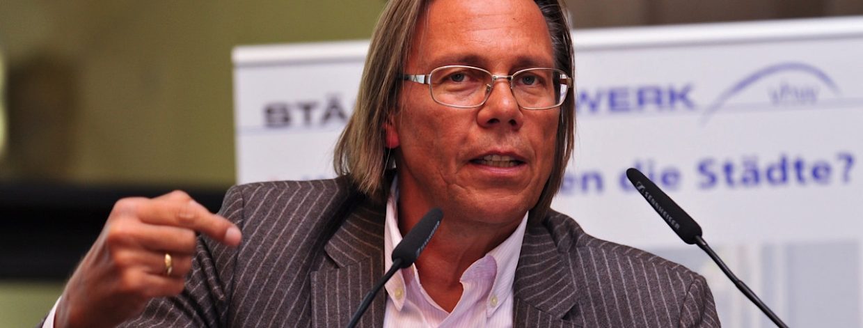 Langhaariger Mann auf Podium vor Mikrofonen