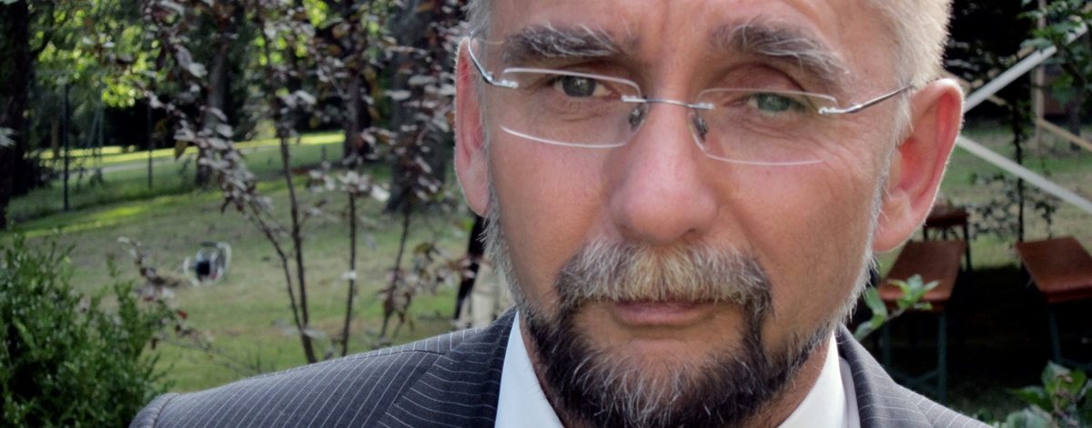 Zynisch blickender Mann mittleren Alters mit leichter Glatze, rahmenloser Brille und Bart in Anzug un Fliege