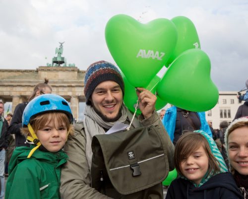 Zwei Kinder, ein Mann und eine Frau stehen vor dem Berliner Tor und halten Herzluftballone in grün mit der Aufschrift AVAAZ