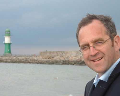 Freundlich lächelnder Mann mit Brille vor Meer und Leuchtturm im Hintergrund