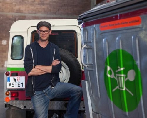 Junger Mann mit Brille und Cappy steht mit verschränkten Armen vor Mülltonne