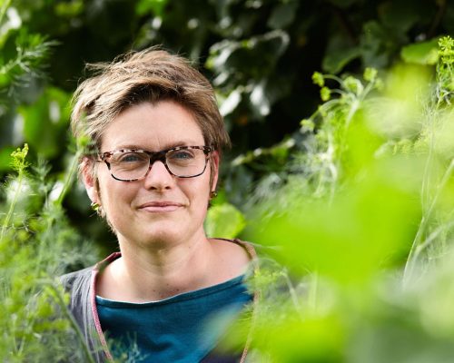 Kurzhaarige Frau mit schwarzer Brille steht in dichten grünen Pflanzen