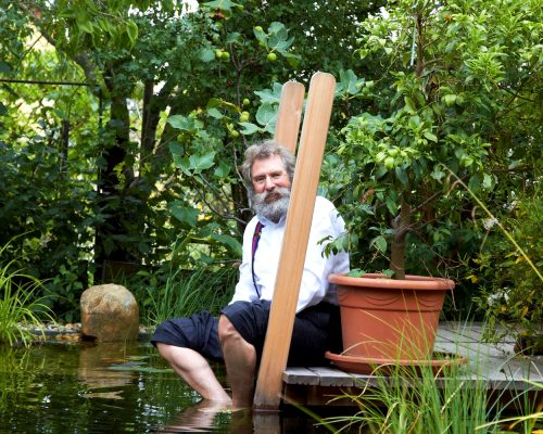 Mann mit grauem Rauschebart und Brill sitzt an Teich mit Füßen im Wasser