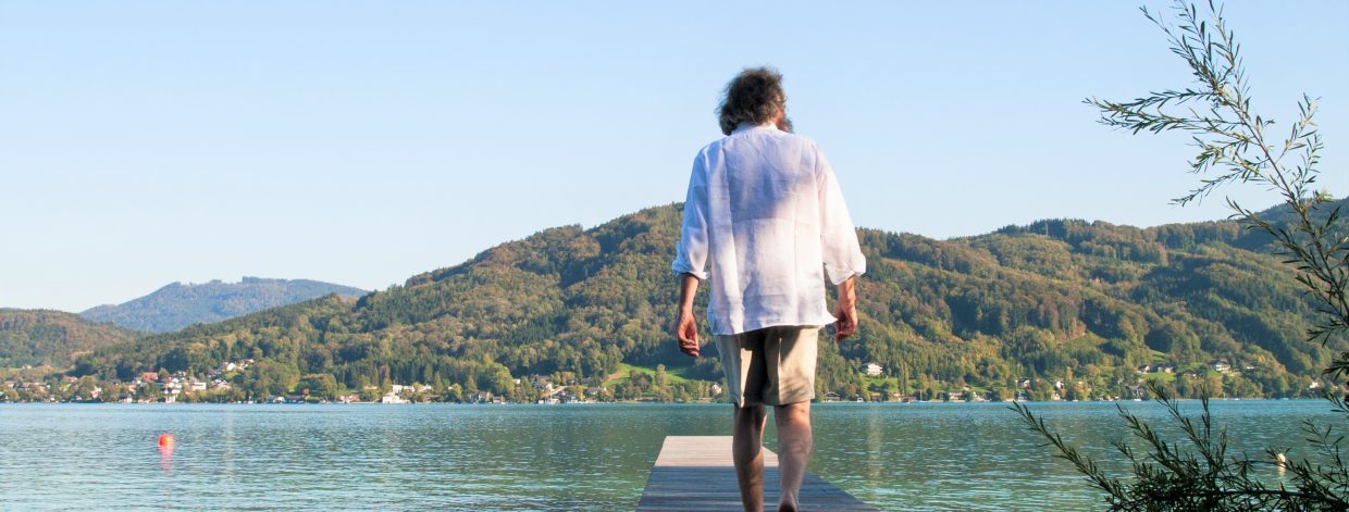 Mann in kurzer Hose und weißem Hemd geht auf einem Steg Richtung See