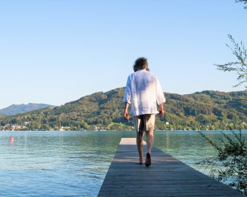 Mann in kurzer Hose und weißem Hemd geht auf einem Steg Richtung See