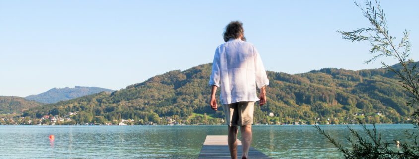 Mann in kurzer Hose und weißem Hemd geht auf einem Steg Richtung See